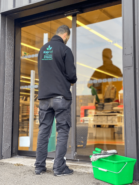 Travailleur de chez Kapnet qui nettoie la vitre d'un magasin avec un sceau vert.