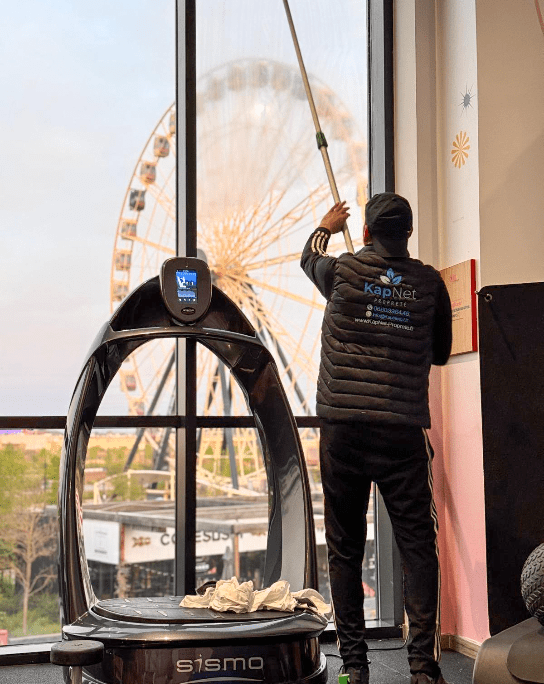 Travailleur de chez Kapnet propreté nettoie une vitre de l'intérieur des locaux. On a vue sur une grande roue.